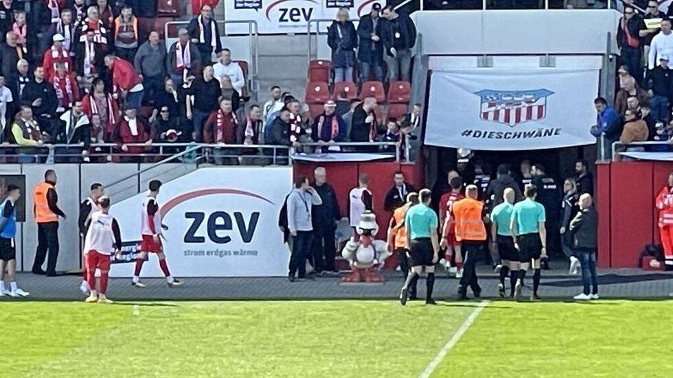 Das Schiedrichter-Gespann auf dem Weg in die Kabine. Der Mann im blauen Pullover wartet rechts oberhalb vom Eingang. 